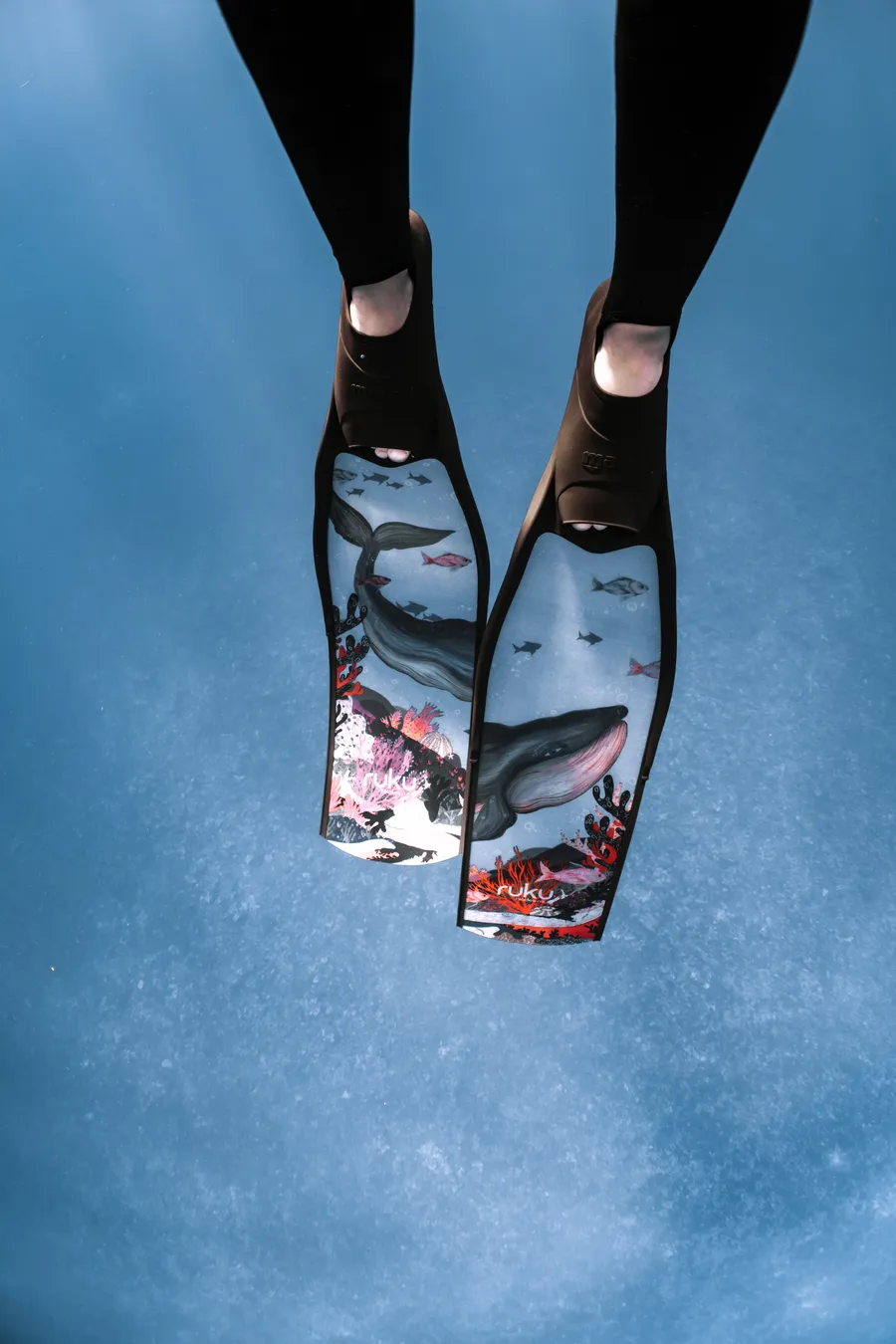 A freediver's painted fins break the silver surface from below — quiet blue abstract